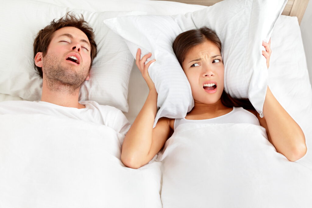 Woman covering her ears with a pillow while lying in bed next to a man who is loudly snoring.